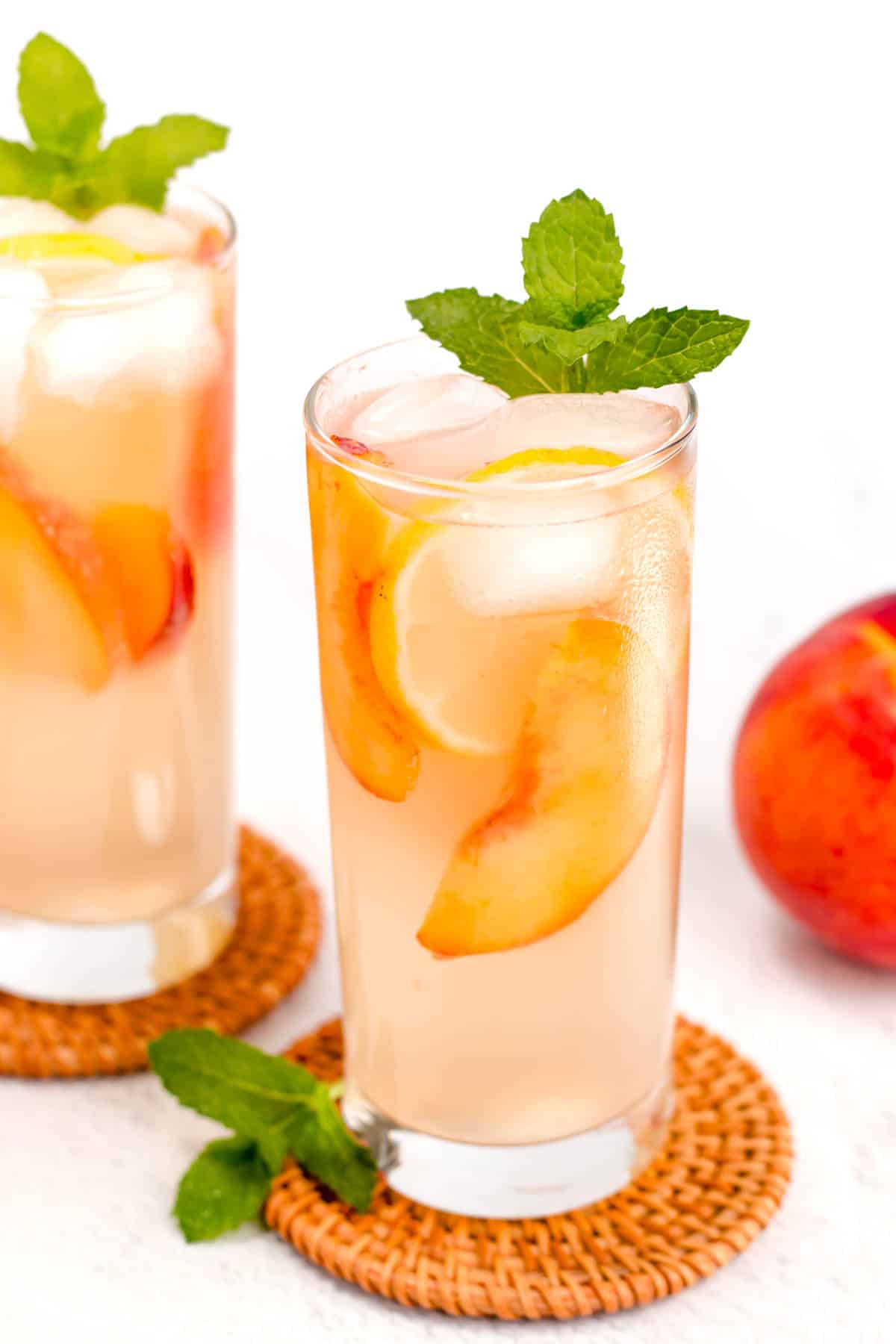 Two tall glasses of iced peach lemonade with lemon slices, peach slices, and mint leaves on top, placed on woven coasters. A fresh peach is visible in the background.