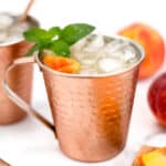 A copper mug filled with an iced beverage garnished with a peach slice and mint leaves, with a metal straw. Another similar mug, peach slices, and a whole peach are in the background.