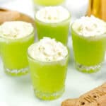 Four neon green drinks topped with whipped cream in small glasses, placed on a white surface with a bone-shaped cookie nearby.