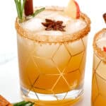 A cocktail in a geometric-patterned glass garnished with a cinnamon stick, star anise, rosemary sprig, and apple slice. The rim is coated with brown sugar and the drink contains ice.