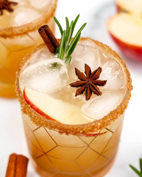 Two glasses of a cold drink garnished with apple slices, star anise, rosemary, and cinnamon sticks, topped with ice and a sugar rim. A partially sliced apple is in the background.