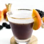 A cocktail in a glass with a slice of peach and a blackberry on the rim, placed on a wooden coaster. There are more peaches and blackberries in the blurred background.