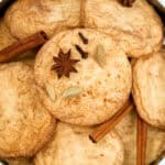 A tin filled with round, lightly browned cookies. The cookies are accented with whole spices, including star anise, cloves, cardamom pods, and cinnamon sticks.