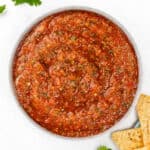A bowl filled with red tomato salsa, garnished with cilantro leaves on the side. Several tortilla chips are placed near the bowl on a white surface.
