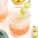 A glass of a light pink drink with crushed ice, garnished with a skewer of green grapes. Another glass and a bowl of green grapes are in the background, with a teal cloth nearby.