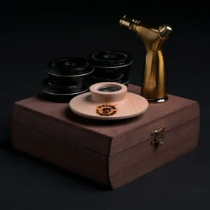 A wooden box with a latch holds stacked black tins, a cocktail smoker, a wooden dish with a metal center, and a gold handheld torch, all arranged on a dark surface.