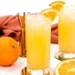 Two glasses of iced orange drinks garnished with orange slices. Fresh oranges and a metal jigger are in the background.
