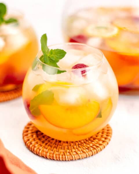 A round glass filled with a fruit-infused drink, ice cubes, and garnished with a sprig of mint sits on a woven coaster. Slices of orange and berries are visible inside the drink.