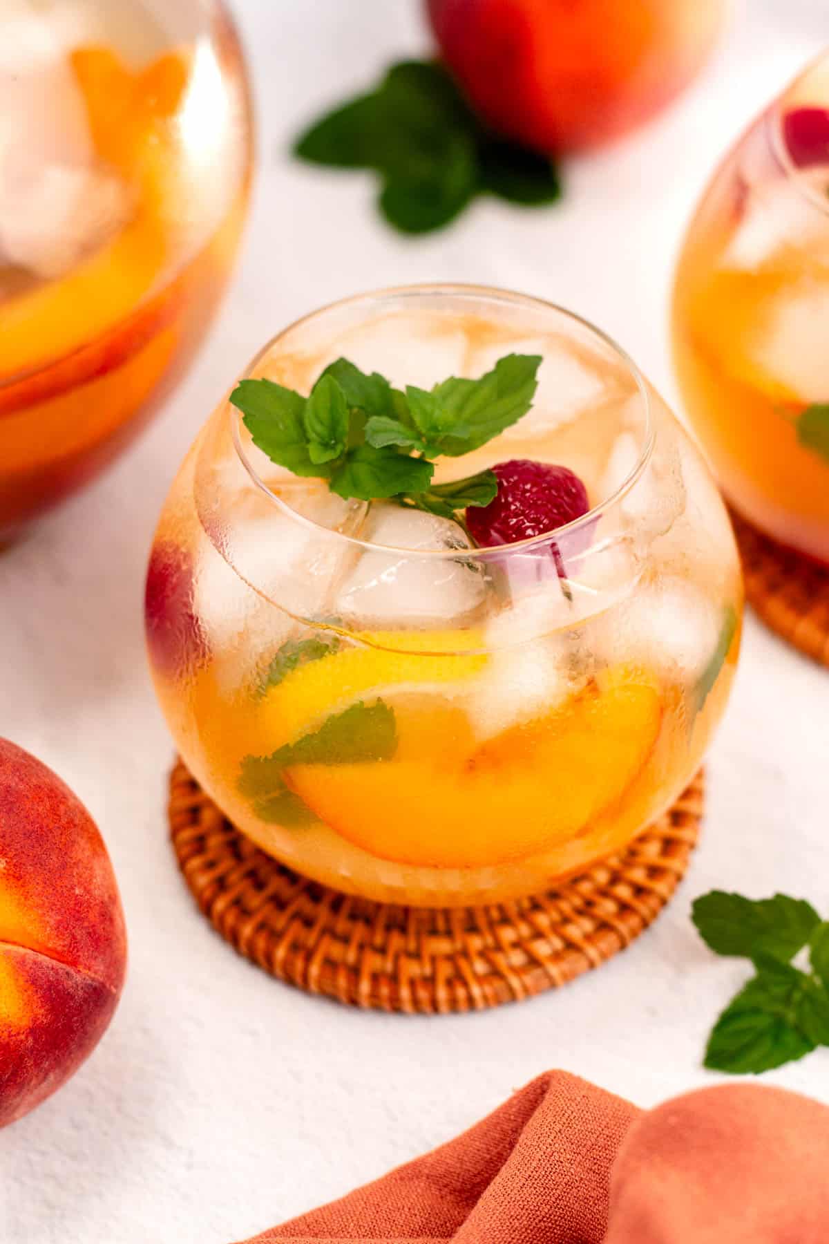 A glass of iced peach sangria is garnished with a mint leaf and a raspberry. Two additional glasses and a fresh peach are visible in the background.