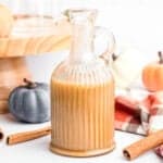 A glass pitcher filled with a beige creamy liquid, surrounded by whole cinnamon sticks, decorative pumpkins, and a plaid cloth, with a wooden stand and glass cup in the background.