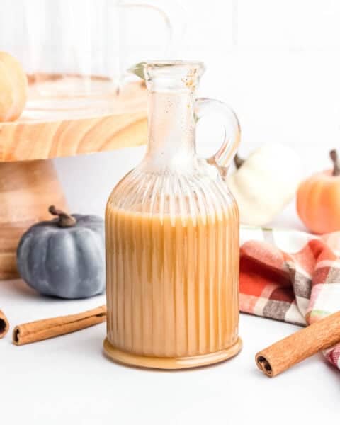 A glass pitcher filled with a beige creamy liquid, surrounded by whole cinnamon sticks, decorative pumpkins, and a plaid cloth, with a wooden stand and glass cup in the background.