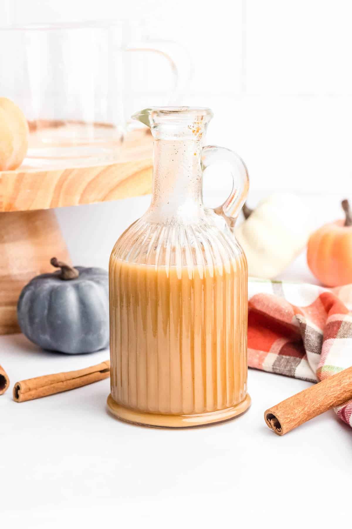 A glass pitcher filled with a beige creamy liquid, surrounded by whole cinnamon sticks, decorative pumpkins, and a plaid cloth, with a wooden stand and glass cup in the background.
