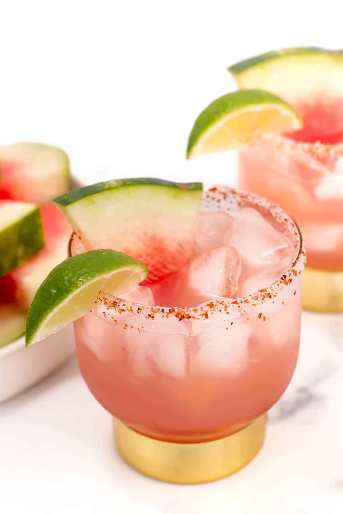 Two glasses of pink-colored cocktails with ice, rimmed with chili powder, and garnished with watermelon slices and lime wedges. A plate with additional watermelon slices is in the background.