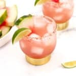 Two glasses of pink cocktail garnished with lime and chili powder on the rim, surrounded by watermelon slices and a jigger on a white surface.