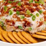 A cheese ball covered in bacon and green onions is placed on a white plate surrounded by circular crackers.