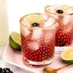 Two glasses of a red iced drink garnished with lime slices and cherries, placed on a pink plate with a spoon, surrounded by halved limes and a bowl of cherries.