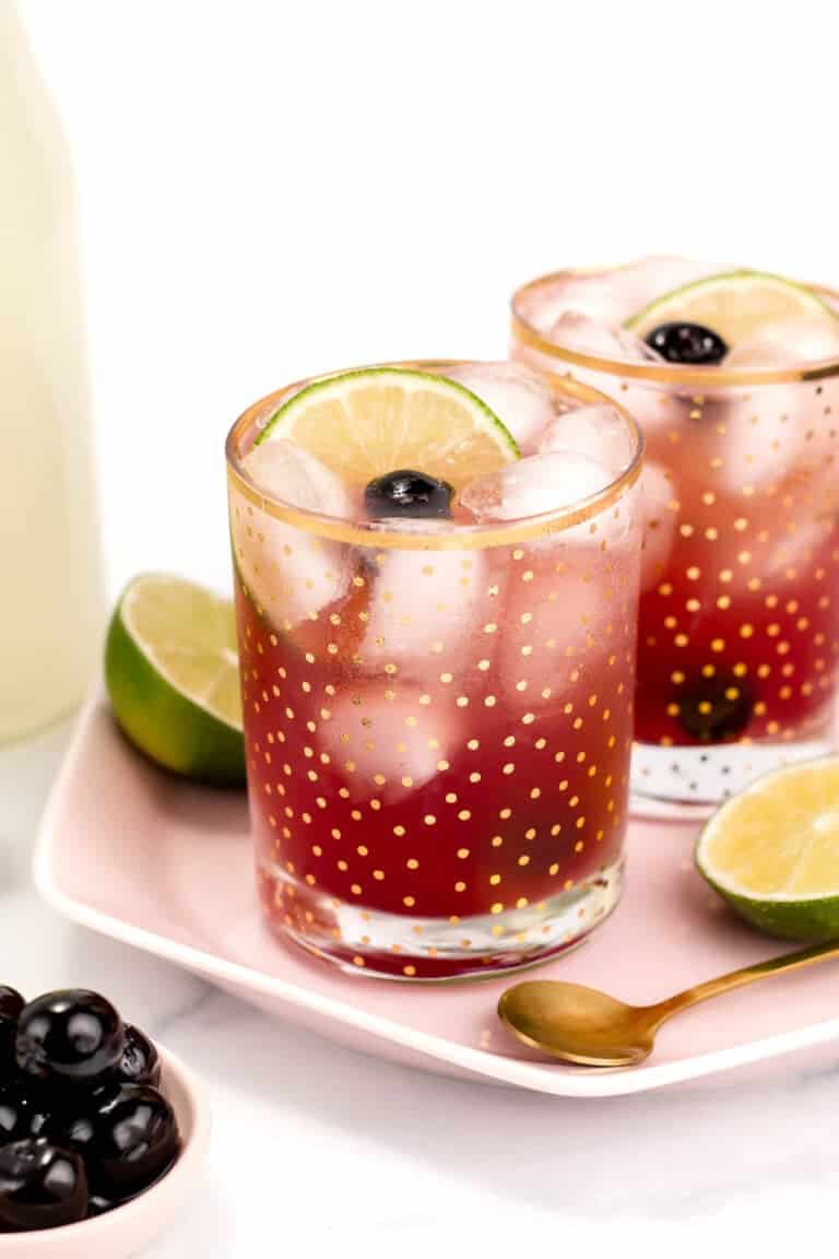 Two glasses of a red iced drink garnished with lime slices and cherries, placed on a pink plate with a spoon, surrounded by halved limes and a bowl of cherries.