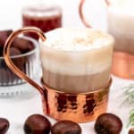 A frothy latte in a copper mug is surrounded by chestnuts and a sprig of rosemary, with blurred jars in the background.