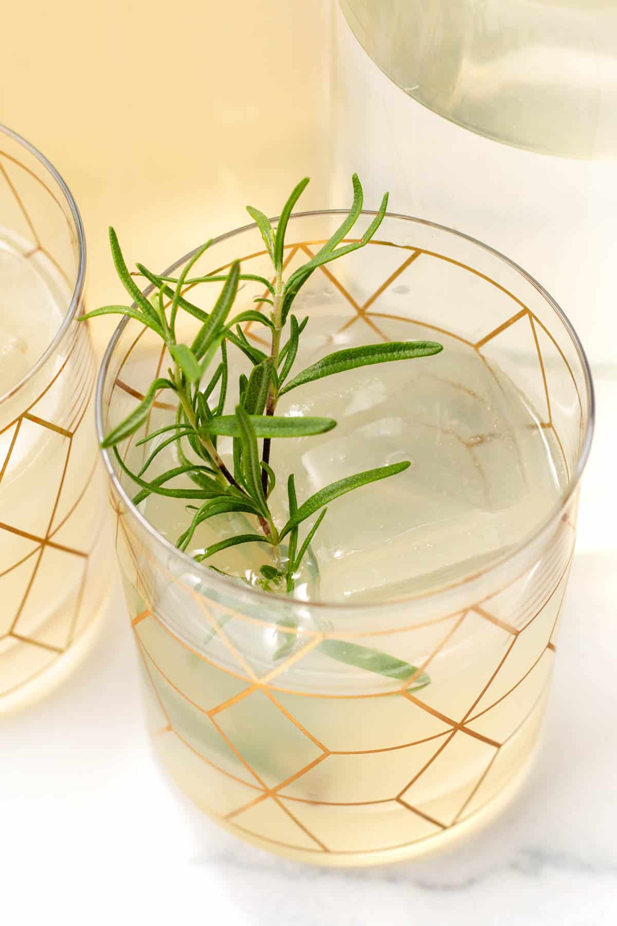 Close-up of a refreshing iced drink in a glass with a gold geometric pattern, garnished with a sprig of rosemary.