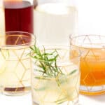 Three geometric-patterned glasses containing different beverages, one garnished with a rosemary sprig, are placed in front of three partially-filled glass bottles.