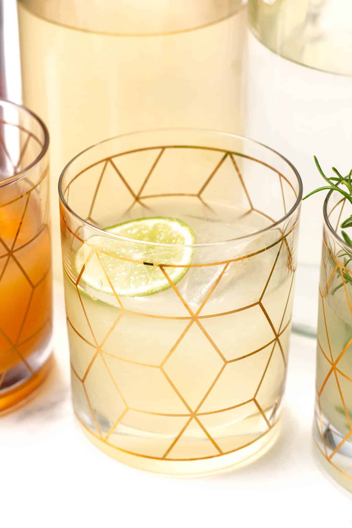 A glass with geometric gold patterns filled with a light-colored drink, garnished with a lime slice. Other beverages in similar decorative glasses are nearby.
