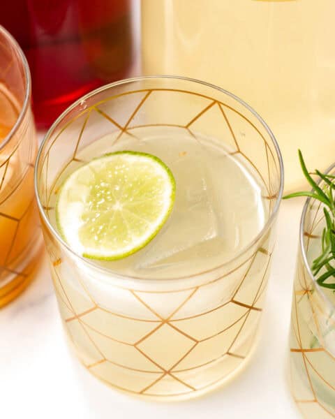 A glass with a geometric pattern contains a light-colored drink, a large ice cube, and a lime slice. Other beverage containers are partially visible in the background.