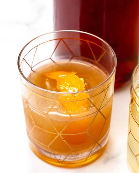 A glass of amber-colored drink with a large ice cube and an orange peel garnish, sitting on a white surface.