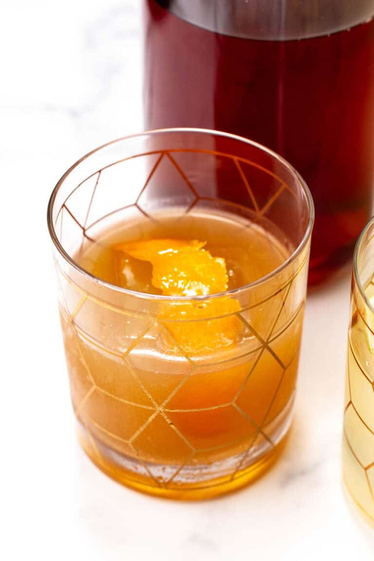 A glass of amber-colored drink with a large ice cube and an orange peel garnish, sitting on a white surface.