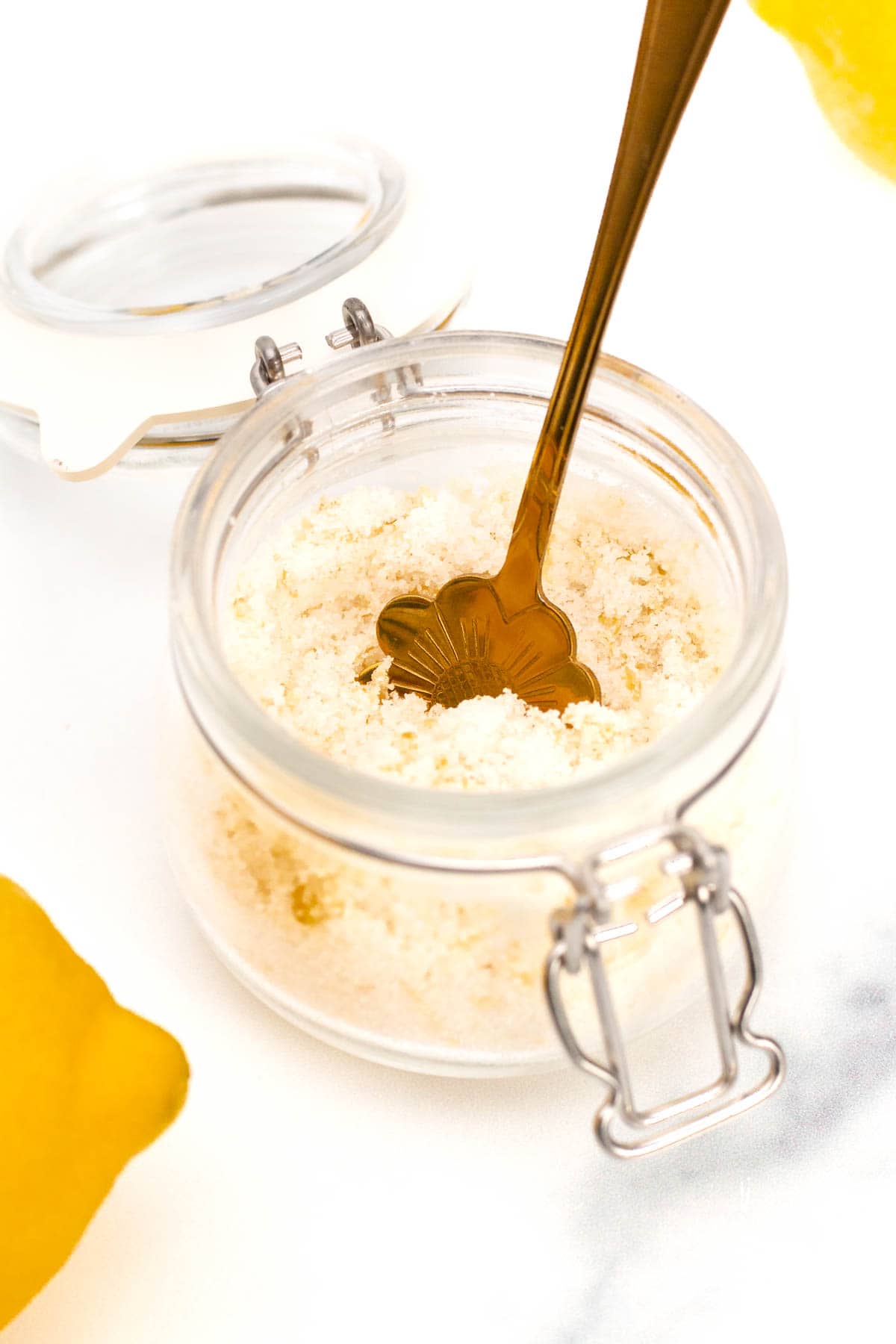 A jar of lemon powder with a gold spoon inside, placed on a white surface, with a cut lemon partially visible in the background.