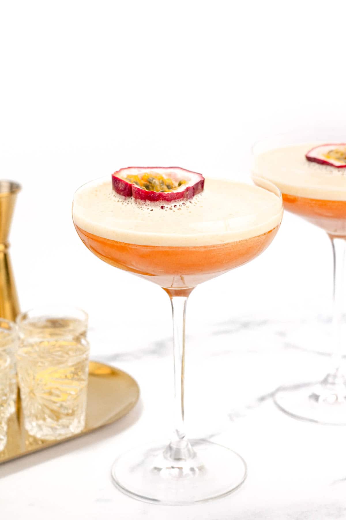 A cocktail glass filled with a foamy orange beverage, garnished with a half slice of passion fruit. A similar cocktail and a tray with liquor glasses are in the background.