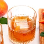 Clear glass with a textured pattern, containing an old fashioned cocktail made with a large ice cube and an orange peel garnish. A peach and a wooden board with peach slices are beside the glass.