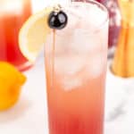 A tall glass filled with an iced, pink to red gradient cocktail topped with a lemon slice and two cherries on a cocktail pick. A lemon, bottle, and bartender tools are in the background.