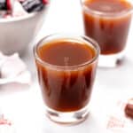 Two small glasses filled with a dark liquid are on a white surface, surrounded by Tootsie Roll candies, some wrapped and some unwrapped, with a bowl of candies partially visible in the background.