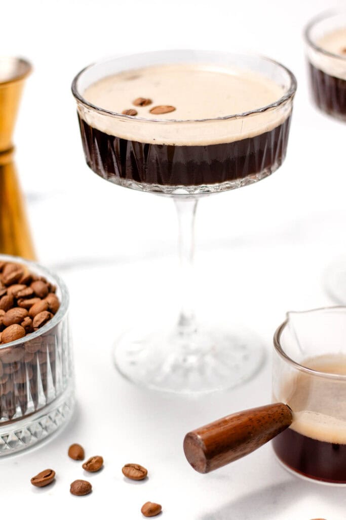 A clear glass filled with a coffee-based beverage topped with foam and coffee beans. Coffee beans and a small pitcher are placed nearby.