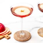 Two coupe glasses of a red cocktail with apple slices, next to sliced apples on a wooden board, against a white background.