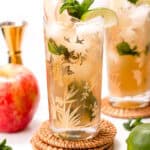 A floral glass with iced apple mojito garnished with mint, cinnamon stick, apple slice, and lime. Served on a woven coaster with a red apple and lime nearby.