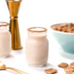 Two small jars of creamy drink with cinnamon rims, next to a gold jigger, a blue bowl of cereal, and scattered cereal pieces on a white background.