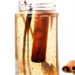 A glass jar filled with light brown liquid, containing cinnamon sticks and vanilla beans. A spoon is dipped into the jar.