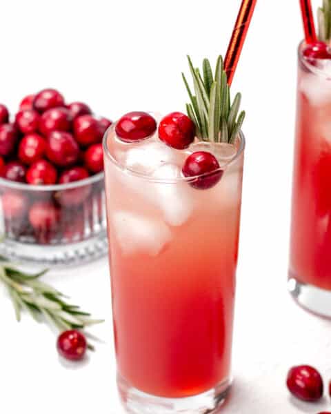A tall glass filled with a pink drink, ice, cranberries, and garnished with rosemary. A bowl of cranberries is in the background.