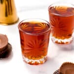 Two etched glass tumblers filled with iced tea sit elegantly on a white surface, accompanied by foil-wrapped chocolate peanut butter cups. It's a perfect setting for a refreshing treat, reminiscent of the indulgence found in a classic peanut butter cup shot.