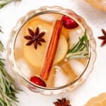 Top view of a glass of spiced apple cider with ice cubes, sliced apple, cinnamon stick, star anise, and rosemary sprigs.