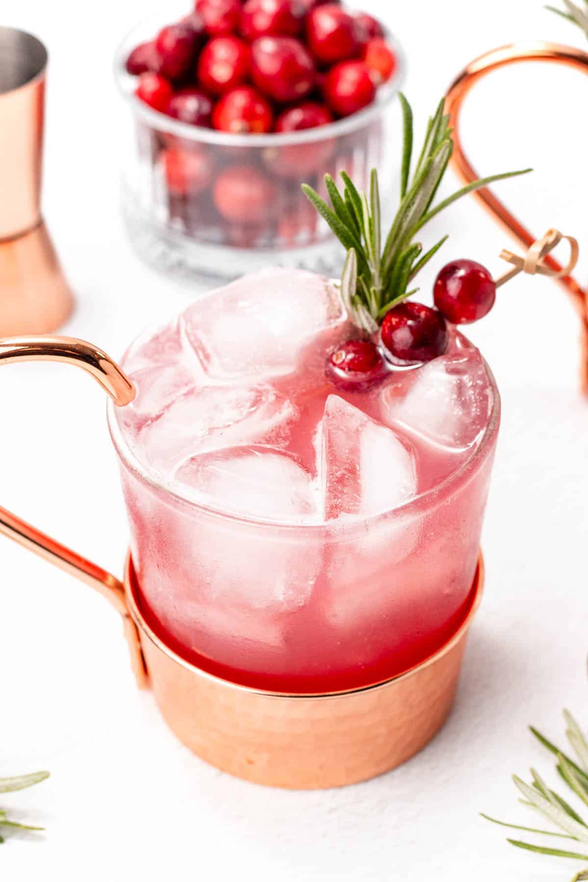 A pink cocktail with ice cubes, garnished with cranberries and rosemary, served in a copper mug. A glass bowl filled with cranberries is visible in the background.