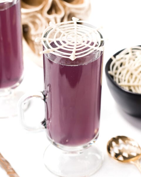 A glass mug filled with a vibrant witches brew cocktail, topped with a white candy spiderweb decoration. A bowl of more spiderwebs and a skull decoration are in the background.