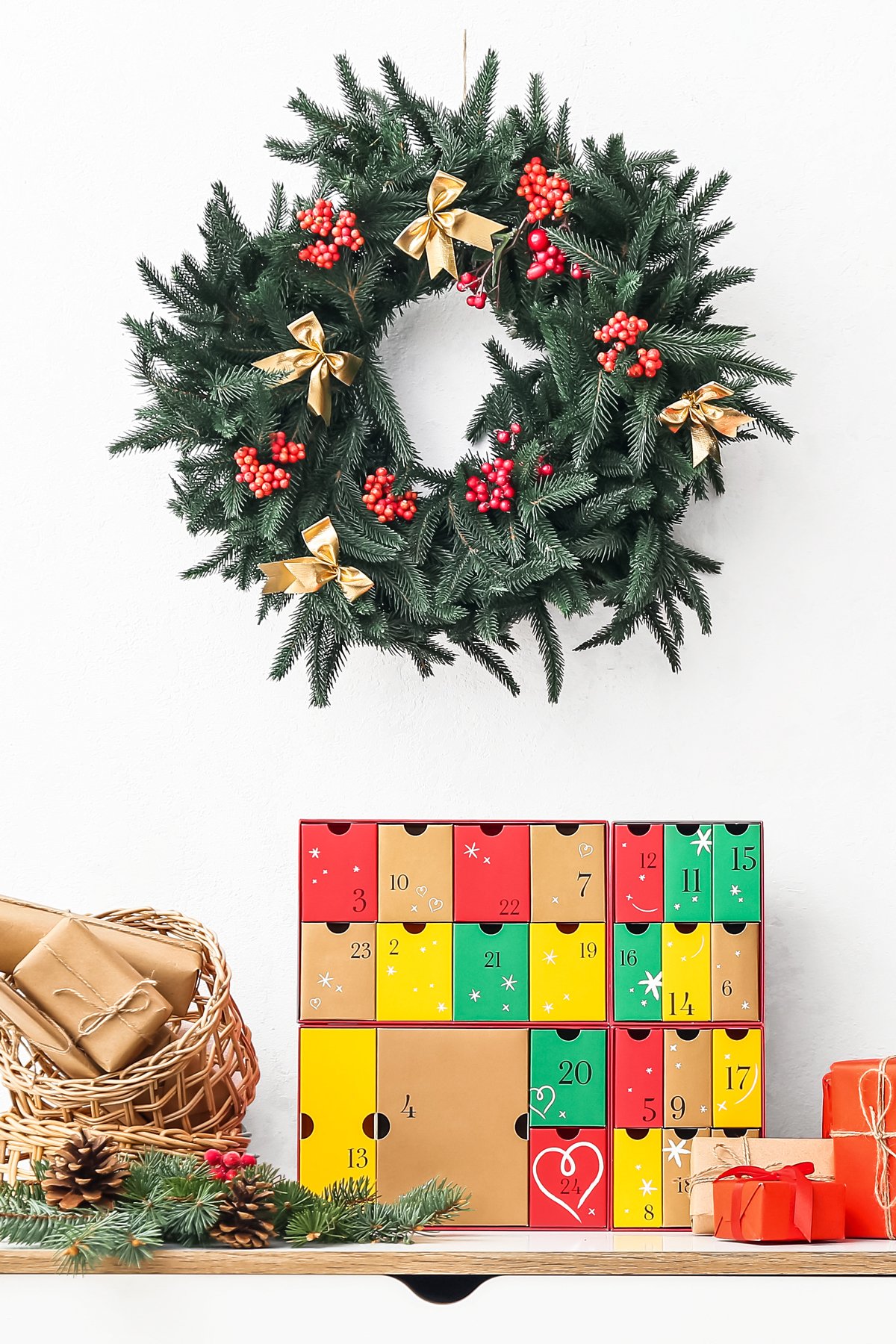 A decorated green wreath hangs above a colorful Food Advent Calendar box, with wrapped gifts, pinecones, and a wicker basket on a wooden shelf below.