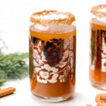 Glass of brown beverage with floral design and sugar rim, reminiscent of a Christmas ale, next to cinnamon sticks and greenery.