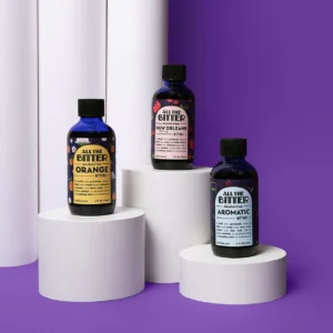 Three bottles from the All The Bitter non-alcoholic bitters bundle—Orange, New Orleans, and Aromatic—are displayed on white cylindrical pedestals against a purple background.
