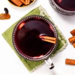 Top view of two cups of mulled wine with cinnamon sticks, surrounded by bowls of raisins and cinnamon sticks on a table.