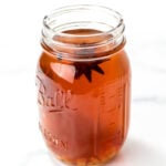 A clear glass mason jar filled with amber-colored liquid, containing visible star anise and small pieces of a light-colored solid, placed on a white surface.