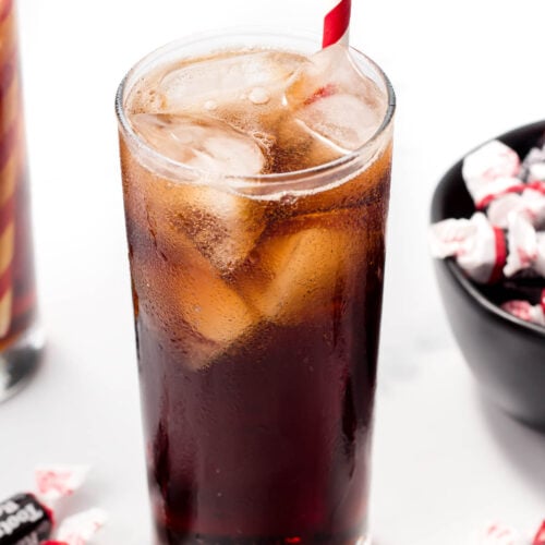 A glass of dark soda with ice and a striped straw is placed on a white surface, surrounded by wrapped candies and a black bowl filled with similar candies.