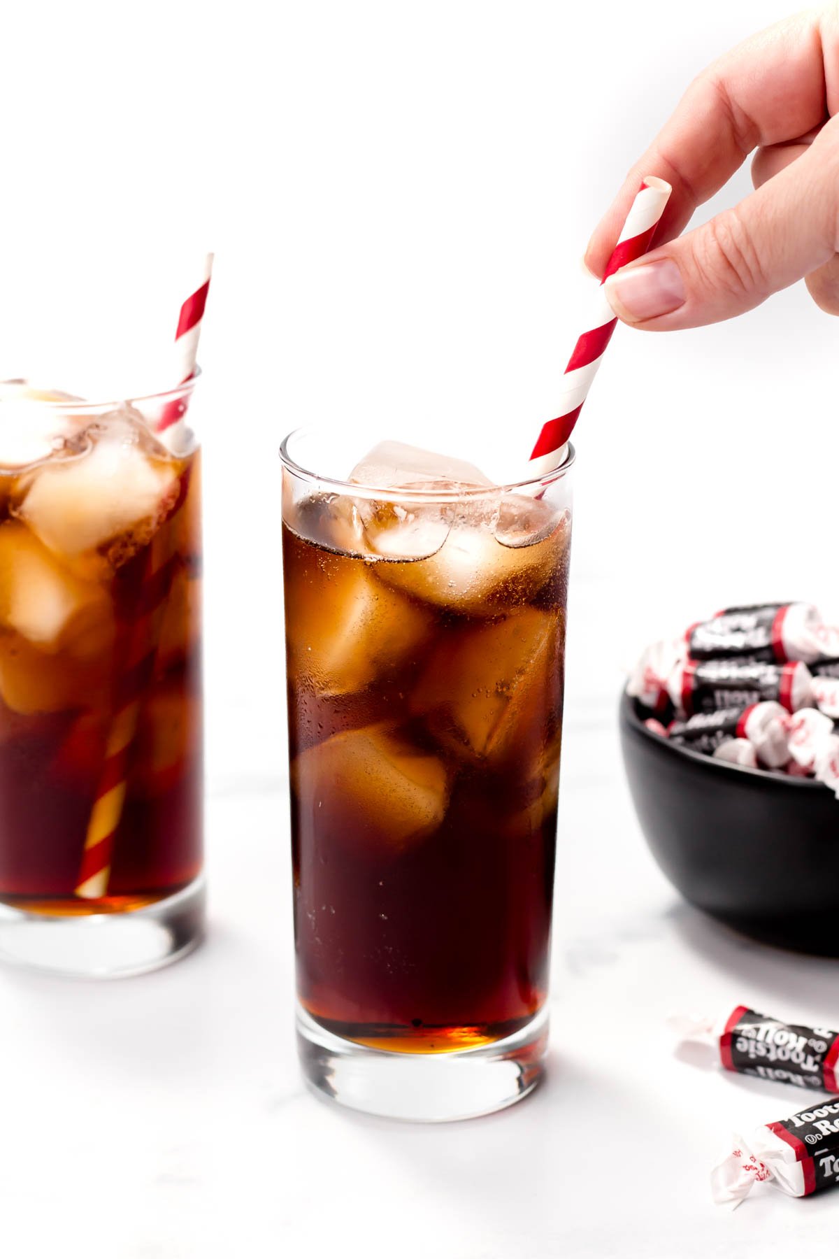 Two glasses of iced cola with striped straws are on a table. A hand adds a straw to one glass. A bowl of wrapped candies is nearby.